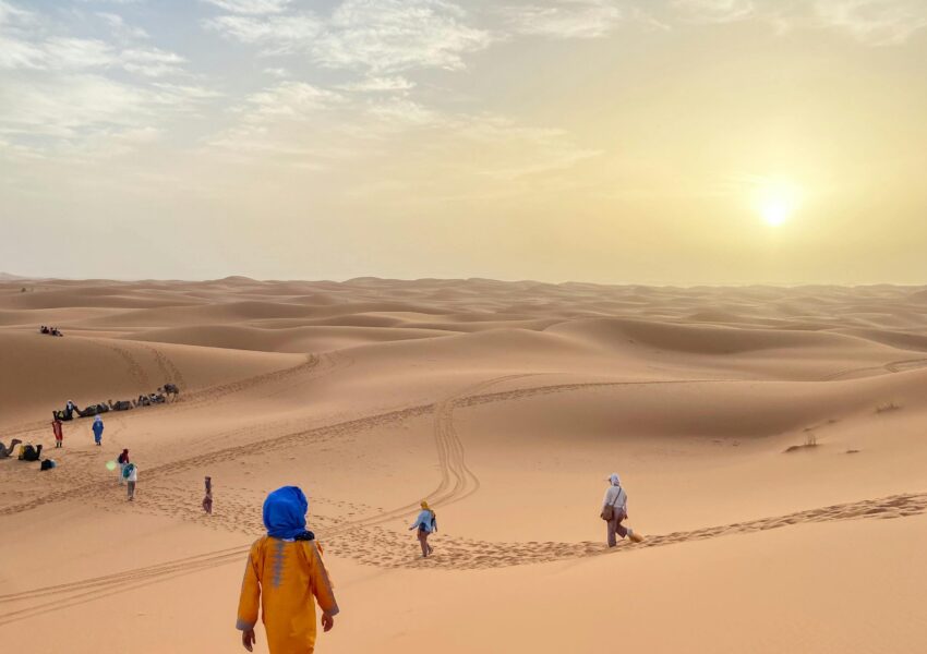 Grupo de pessoas caminhando nas dunas do Deserto de Merzouga ao pôr do sol, com o céu parcialmente nublado.